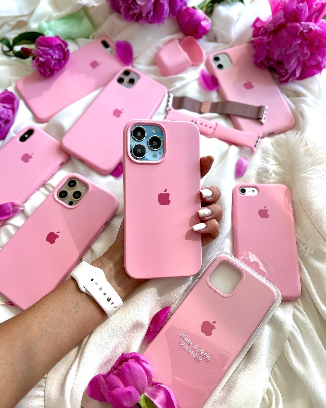 Hand holding an iPhone in a candy pink silicone case among matching cases, accessories, and pink flowers.